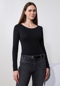 Svart langermet topp med rund hals, parret med mørk grå jeans. Stoffet ser glatt ut, og passformen er tettsittende. Svart belte inkludert.