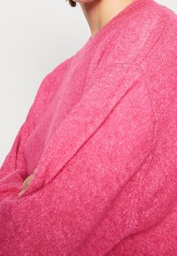 Osoba ubrana w jasnoróżowy, teksturowany sweter z przekroczonymi ramionami, pokazana od ramienia do podbródka na jednolitym tle.