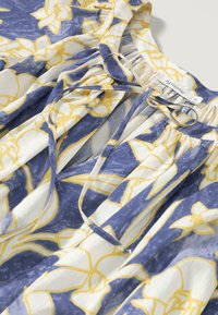 Blumenbluse mit einem blau-weißen Muster und gelben Akzenten. Hergestellt aus leichtem Stoff, verfügt sie über einen gebundenen Ausschnitt und plissierte Details.