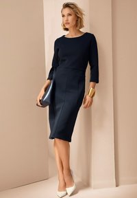 Marineblaues, tailliertes Kleid mit 3/4-Ärmeln, knielanger Saum und vorderer Nahtverarbeitung; kombiniert mit einer kleinen marineblauen Tasche und weißen spitzen Absatzschuhen.