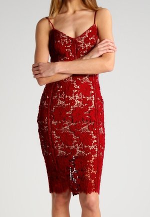 Žena stojící se založenýma rukama na pozadí s jednoduchým vzorem, oblečená v červených krajkových midi šatech bez rukávů s květinovým vzorem.