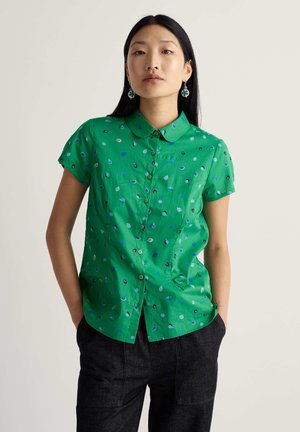 Noor naine, kellel on pikad mustad juuksed, seljas roheline lühikeste varrukatega nööbitav särk sinise lehemustriga ja tumedad püksid, käed taskutes.