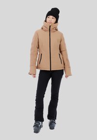 Chaqueta aislante marrón con un cuello alto y bolsillos laterales con cremallera, combinada con pantalones negros y botas de esquí. El atuendo está diseñado para clima frío.