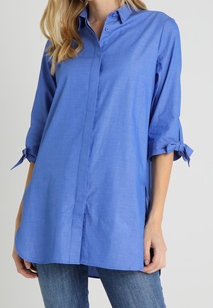 Femme portant une chemise bleue ample à manches longues avec les manches retroussées et nouées en nœuds, associée à un jean bleu, debout devant un fond uni.