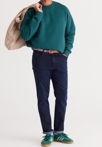 Zielona bluza z okrągłym dekoltem, ciemne jeansy i zielone buty sportowe z białymi paskami. Beżowa torba na ramię zawieszona na jednym ramieniu.