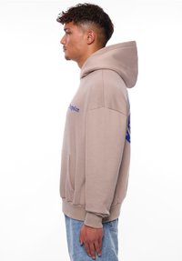 Hellbeige Kapuzenpullover mit großem blauen Grafikdesign auf dem Rücken, Kängurutasche und gerippten Bündchen. Weiche, fleeceartige Textur.