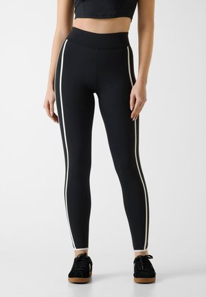 Femme portant des leggings noirs taille haute avec des rayures blanches verticales sur les côtés et des baskets noires avec des semelles marron.