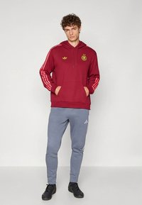 Μπορντό φούτερ με χρυσές λεπτομέρειες και διαμήκεις ρίγες στο πλάι, σε συνδυασμό με γκρι παντελόνι φόρμας με λευκές ρίγες και λογότυπο. Μαύρα αθλητικά παπούτσια.