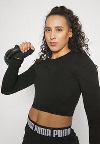 Musthave püsiv mustade pikki varrukatega sportlik tipp, millel on struktureeritud muster ja Puma logo, koos mustade lühikeste pükste ja käes oleva raskusega.