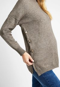 Pull en tricot gris avec des poignets côtelés et trois boutons sur le côté. Équipé d'une fente sur le côté pour plus de facilité et d'une coupe décontractée.