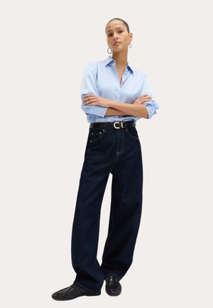Donna in piedi con le braccia incrociate che indossa una camicia azzurra a bottoni, jeans larghi scuri, cintura nera, scarpe nere e orecchini a cerchio.