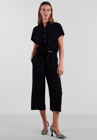 Tuta nera con maniche corte, chiusura a bottoni, vita legata e pantaloni a gamba larga. Abbinata a tacchi a righe bianche e nere.