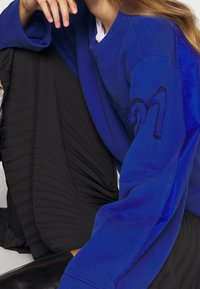 Sudadera azul con detalle bordado "21" en la manga, combinada con una falda negra con pliegues. Tejido suave, ajuste relajado y mangas largas.