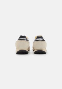 Beige ruskind og sorte syntetiske sneakers med kontrasterende gule detaljer, der har "Saucony"-logoet på hælen og en tekstureret gummisål.