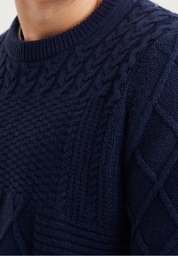 Närbild av en person som bär en marinblå kabelstickad tröja med strukturerade mönster och ribbat rundhals.