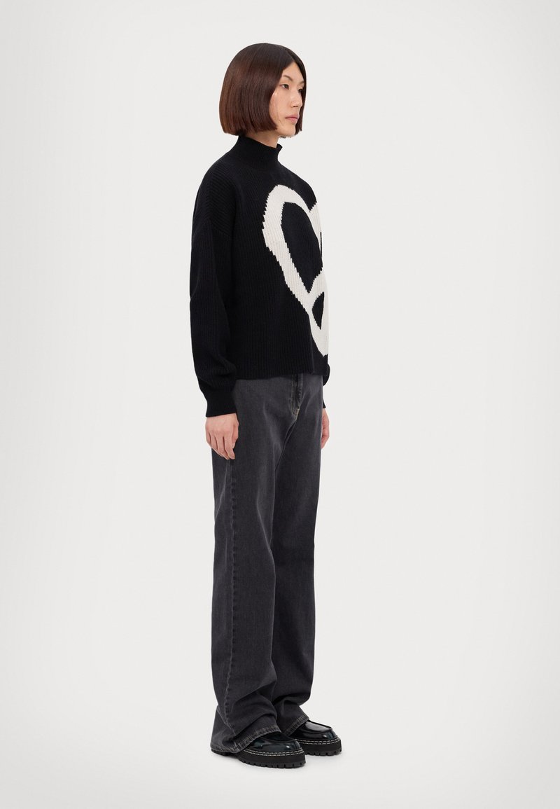 Schwarzer Strickpullover mit weißem Herzmuster, hohem Kragen und lässiger Passform. Kombiniert mit dunkelgrauen weiten Jeans und schwarzen Plateauschuhen.