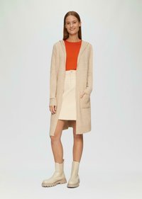 Beżowy długi kardigan z kapturem, noszony na jasnoczerwonym topie, w połączeniu z kremową spódnicą i beżowymi botkami do kostek. Posiada kieszenie i ściągacze na rękawach.