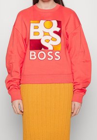Felpa color corallo con vestibilità oversize, caratterizzata da un audace logo "BOSS" in giallo e borgogna. Polsini e orlo a coste, abbinata a una gonna a coste gialla.