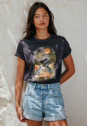 Femme aux longs cheveux foncés portant un t-shirt noir avec un motif de zèbres et de palmiers et un short en jean retroussé devant un mur texturé.