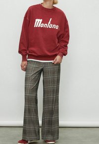 Rode trui met "Montana"-print, gecombineerd met geruite wijde broek in grijs, wit en bruin. Model draagt rode schoenen, minimalistische achtergrond.