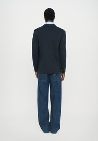 Americana azul marino con un cuello con muescas, combinada con jeans de mezclilla azul de pierna ancha que presentan bolsillos parche y costuras decorativas, mostrada desde la parte trasera.
