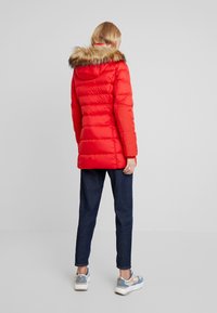 Rød puffjakke med en pelsforet hætte, quiltet tekstur og et mellemlangt design, parret med mørke jeans og lyse sneakers.