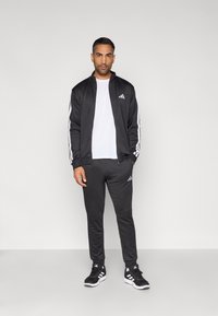 Čierna športová bunda s bielymi pásmi, doplnená o zodpovedajúce tepláky a biele tričko. Má vrecká so zipsom a logá ako akcenty.