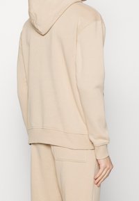 Sudadera con capucha beige de material suave con un bolsillo tipo canguro y puños acanalados. Cuenta con capucha y un ajuste relajado, vista desde atrás.