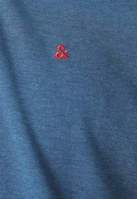 Blå tekstureret stof med et lille rødt broderet ampersand-symbol. Stoffet ser blødt og let vævet ud.