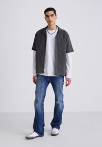 Camisa gris de manga corta sobre una camiseta blanca de manga larga, combinada con jeans azules y zapatillas blancas, de pie sobre un fondo neutro.