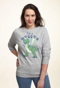 Grå sweatshirt med en grön dinosaurie-grafik och texten "Jag är en KRAMARE" i blått. Tyget har en slät textur och ribbade ärmslut.