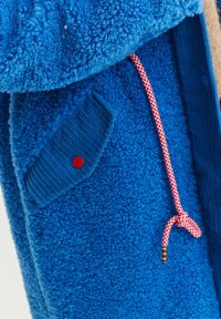 Fleece bunda v žiarivej modrej farbe, s textúrovanou tkaninou, rebrovanými detailmi, diagonálnou vreckom s červeným gombíkom a ružovo-bielym šnúrkami.