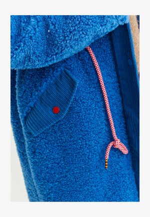 Kurtka polarowa w intensywnym niebieskim kolorze, z teksturowanym materiałem, prążkowanymi akcentami, diagonalną kieszenią z czerwoną guzikiem oraz różowo-białym sznurkiem.