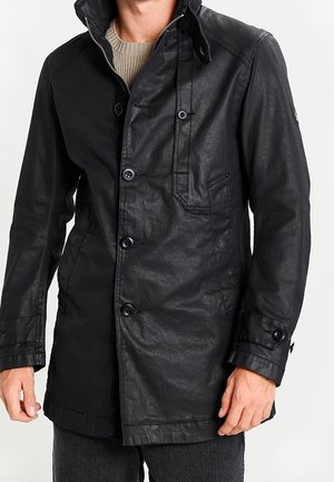 Homme portant une veste en cuir noire boutonnée sur un pull en maille beige et un pantalon foncé, debout devant un fond uni.