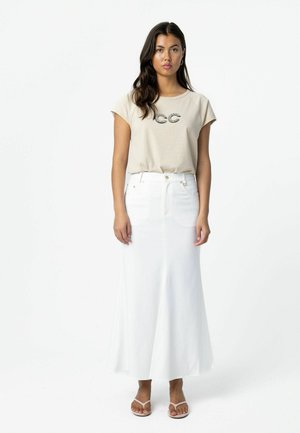 Vrouw met lang donker haar, gekleed in een beige kortermig shirt met "CC"-ontwerp, een witte hooggetailleerde lange rok en witte sandalen, staand tegen een witte achtergrond.