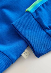 Bluză din bumbac albastră cu accente dungi verzi și galbene. Are manșete reiate și o mică etichetă țesută cu inscripția „Little Bird”.