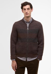 Brown textured knit zip-up sweater with a ribbed collar, and contrasting horizontal stripes. Worn over a checkered shirt and brown pants.