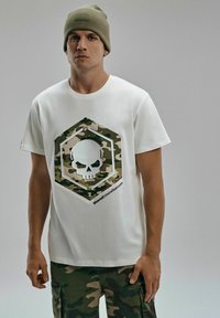 Biała koszulka z dużą heksagonalną grafiką w kamuflażu z czaszką. Model nosi oliwkowe spodnie w kamuflażu i pasującą czapkę beanie.