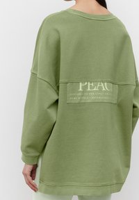 Felpa oversize verde chiaro con collo rotondo e maniche lunghe, caratterizzata da un pannello testurizzato con la scritta "PEACE" e dettagli del marchio sulla schiena.