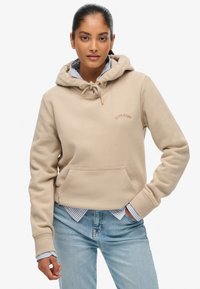 Felpa beige con tasca frontale e cappuccio con cordino. Presenta un logo ricamato ed è sovrapposta a una camicia a righe blu e bianche.