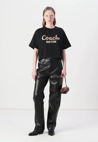 Juodas trumpas marškinėliai su "Coach New York" logotipu, derinami su blizgiais juodais odiniais kelnais, juodais aukštakulniais batais ir maža ruda rankine.