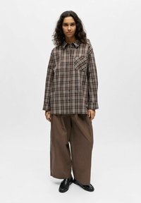 Chemise à carreaux brune avec col boutonné, coupe oversize, poche poitrine, associée à un pantalon large marron et des chaussures noires à enfiler.