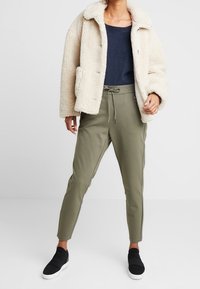 Veste en fausse fourrure crème avec col plat, pull bleu marine, et pantalon olive fuselé avec des bandes latérales. Chaussures noires sans lacets complètent la tenue.