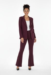Bordowy garnitur z dopasowaną marynarką zapinaną na dwa guziki i kieszeniami z patkami. Dopasowane spodnie z rozszerzanym dołem, zestawione z białą bluzką.