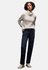 Jersey beige de punto con cuello vuelto, textura estampada; combinado con vaqueros rectos de pierna en azul marino oscuro y zapatillas de deporte de color claro.