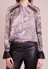 Blusa transparente de manga larga con estampado de pájaros, puños de encaje y detalle de lazo en el cuello, combinada con pantalones de cuero negros.