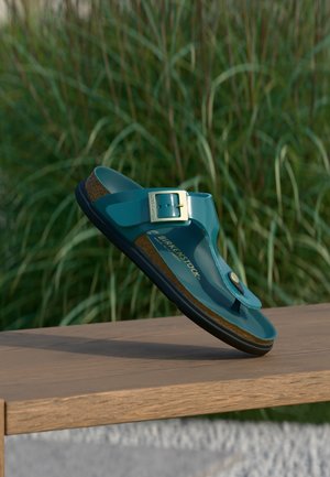 Blå Birkenstock-sandal med spænde og tårem over en træbænk med grønne planter i baggrunden.