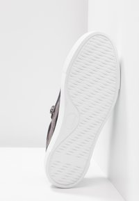 Chaussure grise avec semelle en caoutchouc blanche texturée reposant contre une surface blanche d'angle, montrant le motif complet de la semelle.