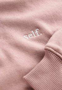 Felpa rosa chiaro realizzata in un tessuto morbido, con polsini a coste e logo ricamato "self." in bianco.