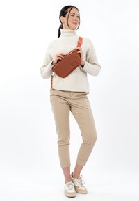 Bruine crossbodytas met structuur, gemaakt van synthetisch materiaal, voorzien van een ritssluiting en verstelbare riem. Draagt een witte trui en beige broek.